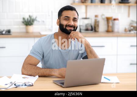 Portrait of handsome positive young adult Indian or Arabian man, freelancer, designer or IT specialist, sit in the kitchen, works from home using laptop, looking directly into camera, smiling friendly Stock Photo