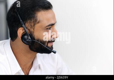 Side view of attractive indian or arabian, call center operator, hotlane expert, manager, uses headset for conference calling, conducts distant consultation, looking at the camera and smiling friendly Stock Photo