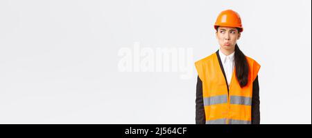 Gloomy and offended young asian female construction manager, industrial woman in safety helmet complaining or whining, pouting sad and looking upper Stock Photo