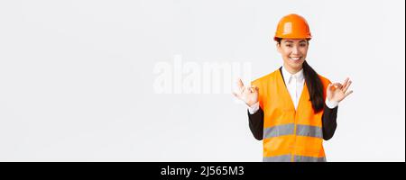 Architect made perfect plan. Confident asian female engineer in safety helmet showing okay gesture, have all under control, pleased with excellent Stock Photo