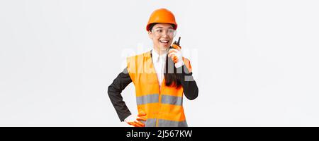 Confident pleased asian female engineer in safety helmet and uniform talking with chief architect using walkie-talkie. Satisfied construction Stock Photo