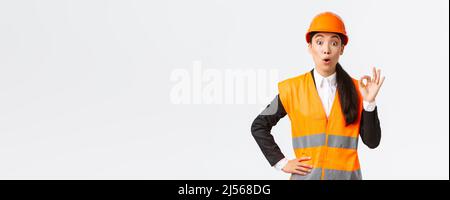 Imressed and surprised asian female construction manager pleased with amazing quality, standing in safety helmet and reflective jacket, showing okay Stock Photo