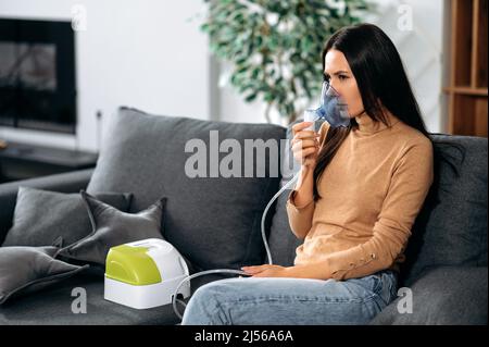 Ill caucasian young woman with an inhaler, sick upset girl doing inhalation at home, she use nebulizer and inhaler for the treatment while sitting on the couch at home at quarantine Stock Photo