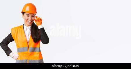 Confident successful asian female chief manager looking proud and smiling at camera, wearing safety helmet, glasses and gloves at construction area or Stock Photo