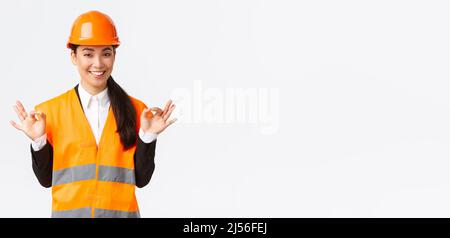 Architect made perfect plan. Confident asian female engineer in safety helmet showing okay gesture, have all under control, pleased with excellent Stock Photo