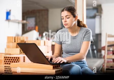 Woman designer sitting on brick stack in construction site and using laptop Stock Photo