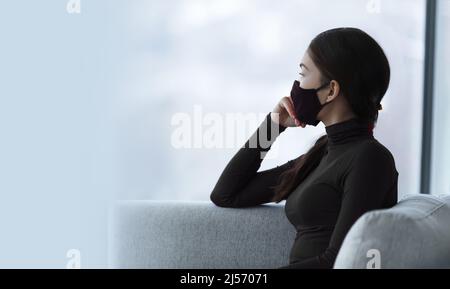 Mask wearing protection from Covid-19 coronavirus. Sad Asian woman wearing face mask feeling depressed alone at home pensive looking at window Stock Photo