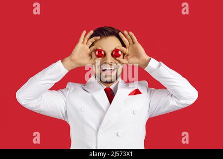 Handsome lucky man smiling and covering both his eyes with two red poker chips in hands, standing on red studio background, copyspace . Casino and Stock Photo