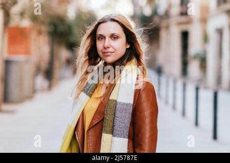 Charming female in leather jacket and scarf looking at camera while standing on sidewalk in city with buildings on blurred background Stock Photo