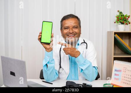 Smiling doctor showing green screen mobile phone by pointing finger while looking at camera at hospital - concept of mhealth, virtual health Stock Photo