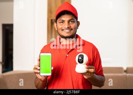 happy technician showing green screen mobile phone by holding smart cctv camera while looking camera at home - concept of technology, advertisement Stock Photo