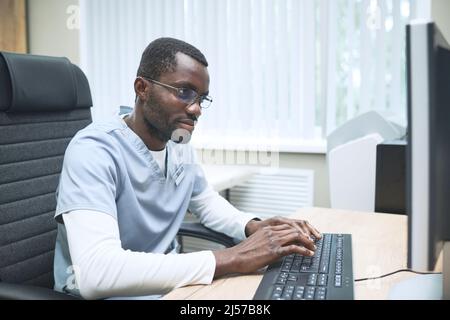 Busy young Black doctor with stubble sitting at desk in office and typing on computer Stock Photo