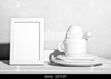 white tableware and empty frame on wooden table. Dishes ans tea cup stacked next to white frame mockup. copy space. traditional dinnerware , kitchen cleaning concept Stock Photo