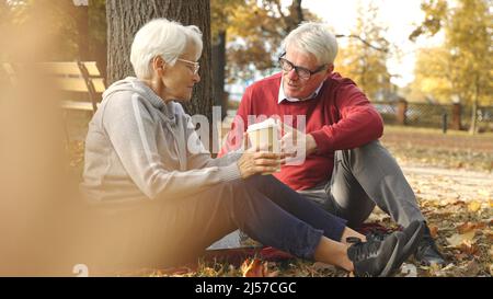 Elderly pensioner lady and her gray-hair husband spend time together in park, sitting on a blanket, drinking coffee, and casually talking. Autumn vibes. High quality photo Stock Photo