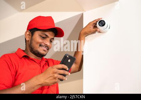 Smiling cctv technician busy using mobile phone for check settings or controlling at home while on ladder - concept of safety, technology and Stock Photo