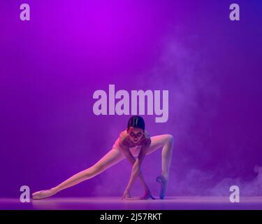 Beautiful flexible young female ballet dancer, teen in stage outfit and pointes dancing isolated on purple background in neon light with smoke. Art Stock Photo