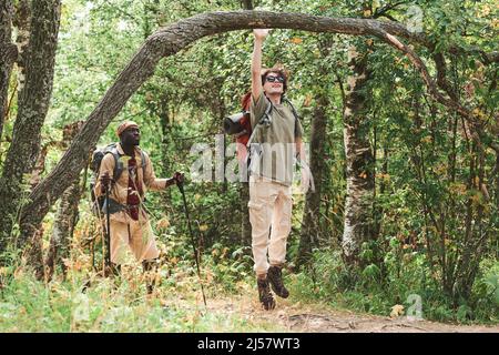 Positive young guy in sunglasss jumping to tree trunk while walking with black friend in forest Stock Photo