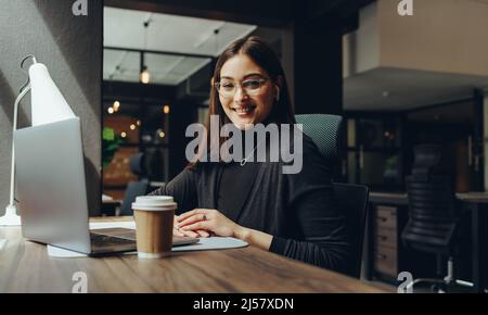 Cheerful young businesswoman smiling at the camera while working in a modern co-working office. Happy female entrepreneur using wireless earphones and Stock Photo