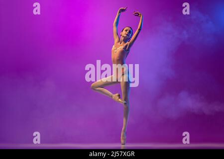 Beautiful flexible young female ballet dancer, teen in stage outfit and pointes dancing isolated on purple background in neon light with smoke. Art Stock Photo