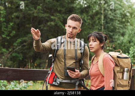 Young Caucasian hiker with camera hanging on neck gesturing hand and using phone while explaining route to woman Stock Photo