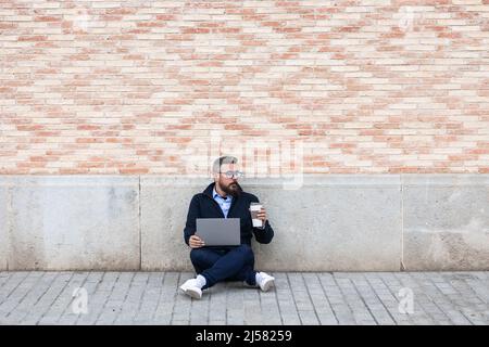 Relaxed man sitting on floor and leaning against wall using his laptop while holding coffee cup Stock Photo