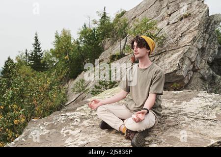 Content relaxed young Caucasian man in hat sitting in lotus position and meditating on big stone outdoors Stock Photo