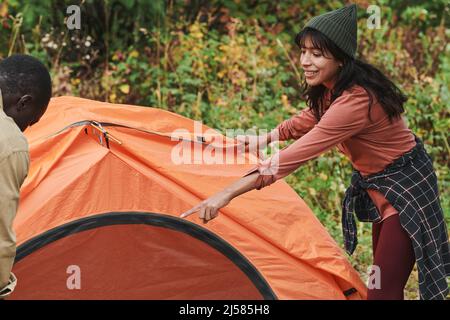Positive young mixed race woman in hat pointing at pole while assisting boyfriend to set up tent at camping Stock Photo