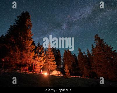 Night camping on mountain hill. Bottom view of two campers sitting opposite each other by the fire next to tourist tent. Gathering couple of friends near bonfire at warm starry evening. Stock Photo