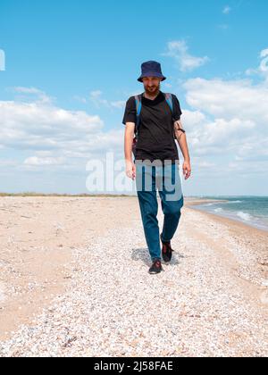 Smiling bearded millennial man in bucket hat black t-shirt walking on empty beach. Authentic male tourist lifestyle photo. Hipster guy outdoor. Solo t Stock Photo