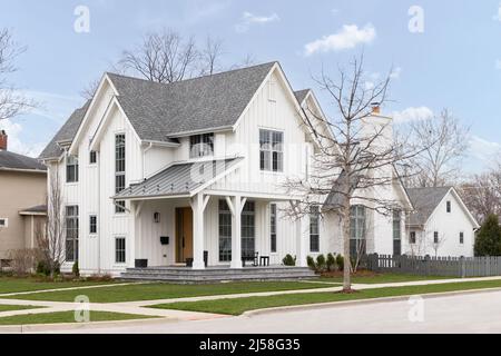 A white modern farmhouse with a covered front porch, board and batten siding, a wooden front door, and grey fence. Stock Photo