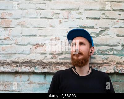 Portrait of young red bearded man in black t-shirt and snapback in rock style on urban background listening to music with headphones. Hipster guy in w Stock Photo