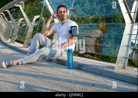 Confident relaxed handsome Caucasian man, athlete in sportswear, putting on headphones and listening to the music while relaxing on the urban glass br Stock Photo