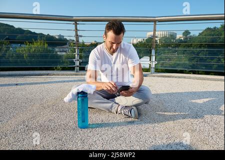 Middle aged Europen man, athlete using fitness tracking app on smartphone. Determined sportsman checking his phone , sitting on the city bridge and re Stock Photo
