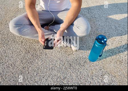 Overhead view of an athlete wearing earphones sitting on the city bridge and checking his phone while relaxing after morning cardio workout and using Stock Photo
