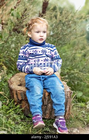 A little girl toddler is sitting on a stump in a spring park. Walk. Stock Photo