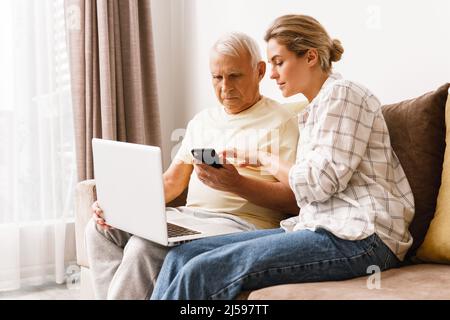Young woman explaining to elderly man how to use laptop and smatphone Stock Photo