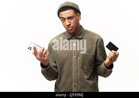 Latin man worrying about money. Confused man holding credit card and smartphone with disappointed face, shrugging, bankruptcy concept Stock Photo