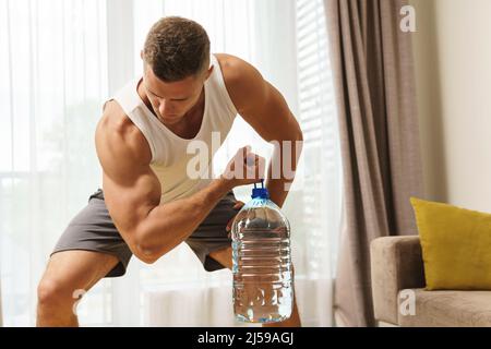 Young athletic man using big bottle of water like an alternative of dumbbell for home workout Stock Photo