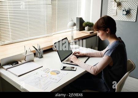 Concentrated young businesswoman sitting at desk and using laptop while communicating with client online Stock Photo