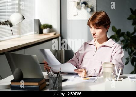 Serious young Caucasian businesswoman with short hair sitting at table and using laptop while doing paperwork Stock Photo