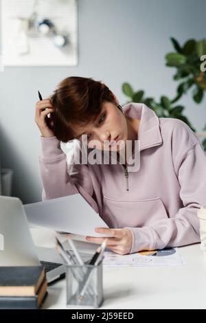 Puzzled young female manager in sweatshirt sitting at desk and reading papers while analyzing data in office Stock Photo