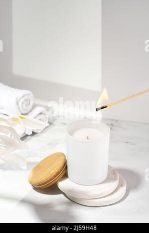 An aromatic soy wax candle is set on fire with match. An ecological and vegan candle, bath accessories and a wooden cap on marble table. Aromatherapy Stock Photo