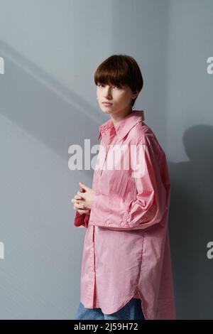 Portrait of serious beautiful young Caucasian woman in long shirt and earrings standing with clasped hands against gray wall Stock Photo