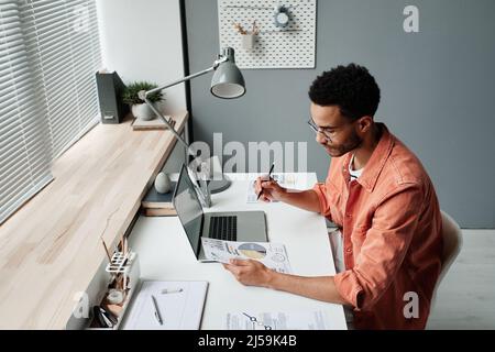 Thoughtful young mixed race man in eyeglasses sitting at table with lamp and analyzing graphs and charts Stock Photo