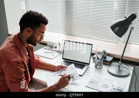 Serious young mixed race project manager with beard sitting at desk with laptop and examining charts in office Stock Photo