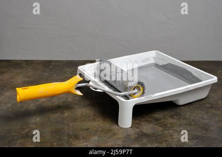 Paint roller with yellow handle in tray for roller with gray paint on grey painted wall background Stock Photo