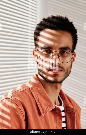 Portrait of serious young black man in round-shaped eyeglasses and coral shirt sitting against closed blinds Stock Photo