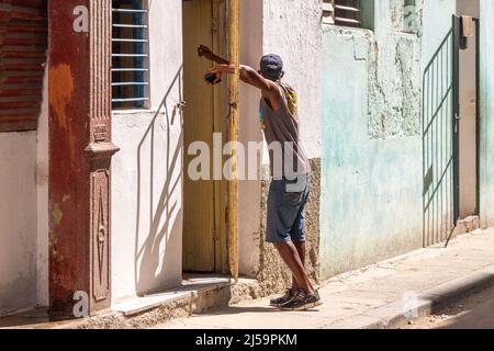 A thin Afro-Caribbean Cuban man is waiting at a house door. He is leaning on a protective metallic grid which is half open. Lifestyle of real people i Stock Photo