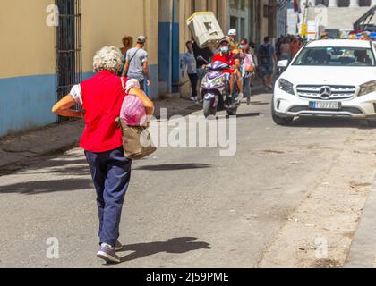 A senior Cuban woman wearing a red blouse walks in a city narrow street where other people are seen in their daily lifestyles. Some are carrying a box Stock Photo