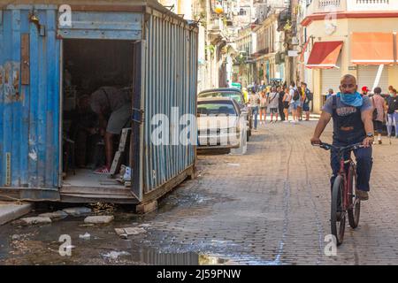 Dirty water is seen by a freight container that is being used as storage. Other Cuban people are seen in their normal routines in the cobblestone stre Stock Photo